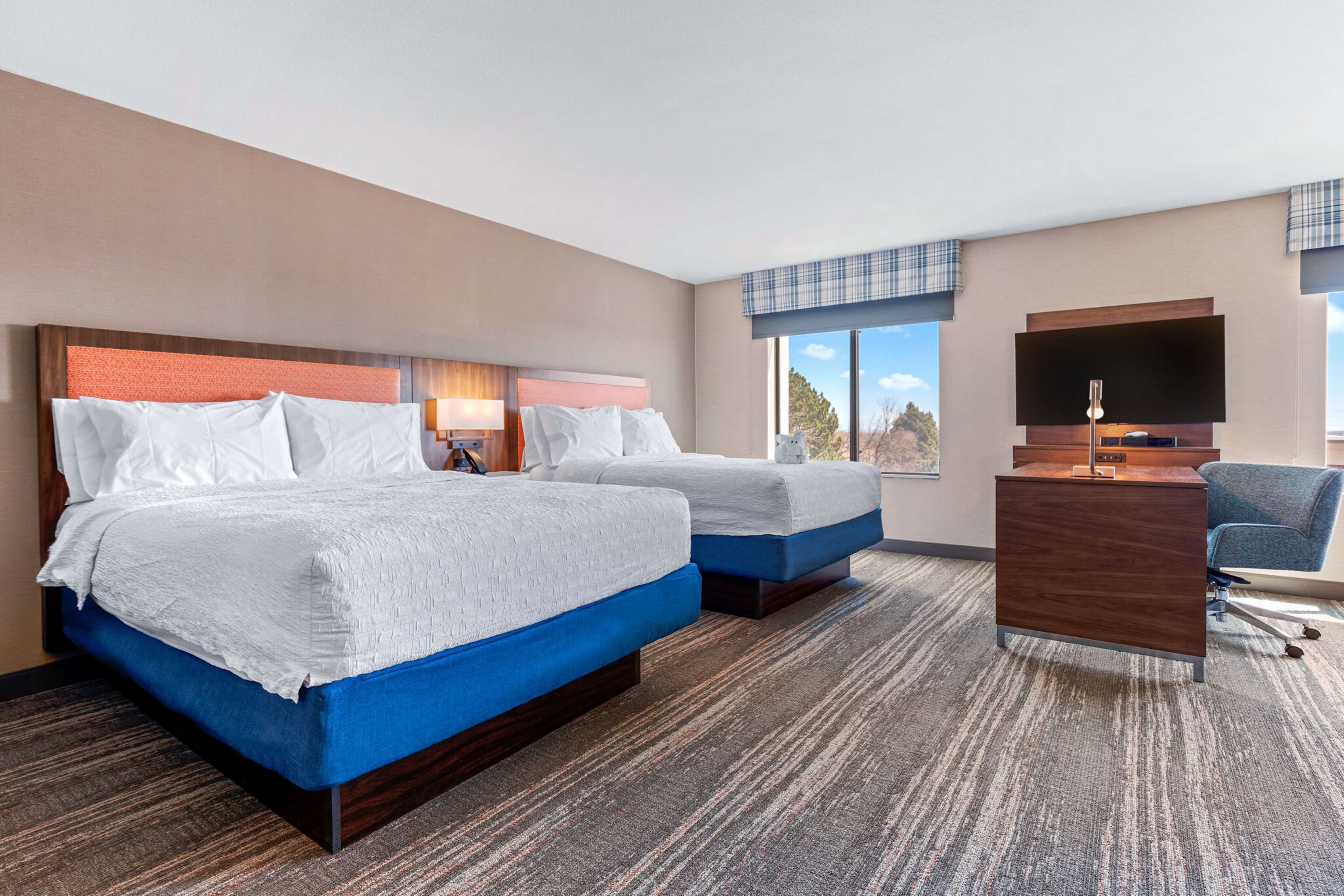 A hotel room with two beds, a desk, a chair, a TV, and a window with a view.