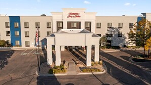 A Hampton Inn & Suites hotel with a central entrance pavilion, parking lot, and a blue building adjacent to it.