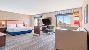 Habitación de hotel con cama, escritorio, silla, televisión y ventanas amplias con vistas.
