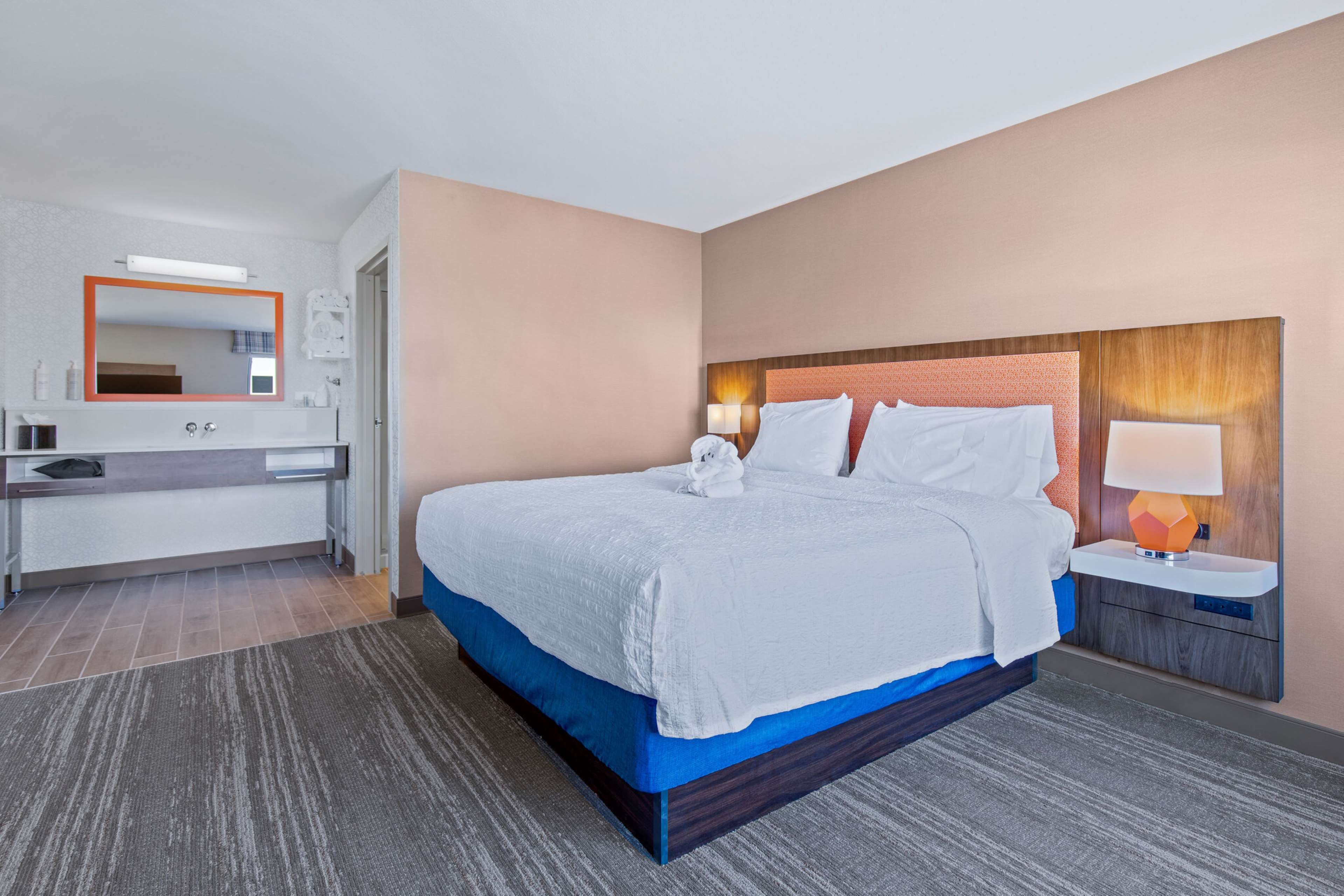 A hotel room with a large bed, a bedside table with a lamp, a mirror above the dresser, and a bathroom with a sink and mirror.