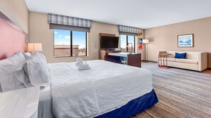 A hotel room with a large bed, a desk, a TV, a sofa, and a window with a city view.