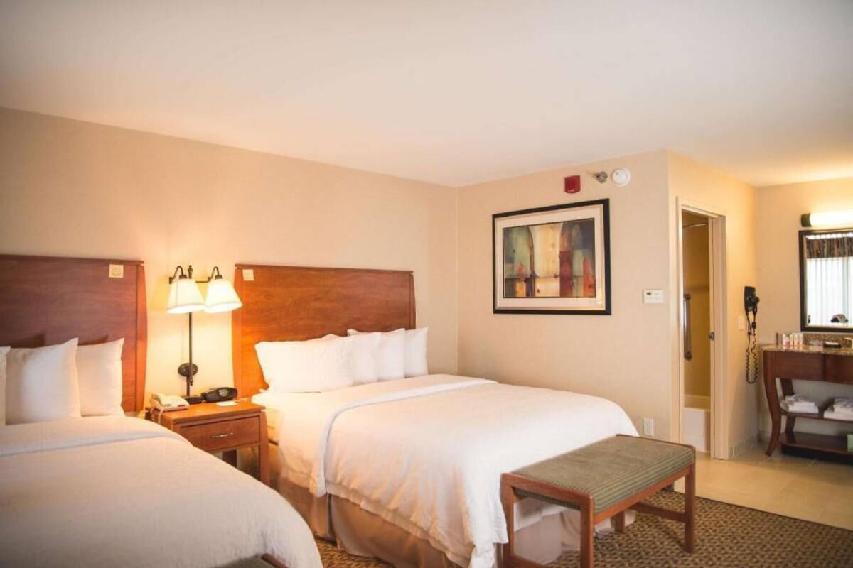 A hotel room with two beds, a bedside table with a lamp, a painting on the wall, a desk with a telephone, and a mirror.