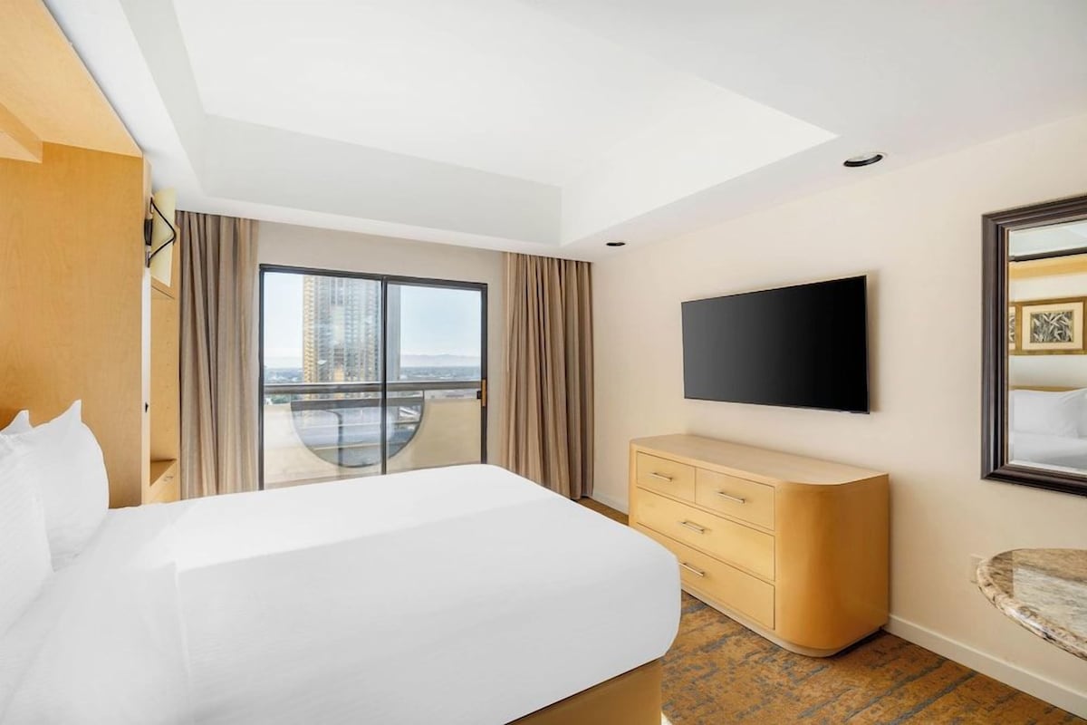 A hotel room with a large bed, a flat-screen TV, a wooden dresser, and a balcony with a city view.