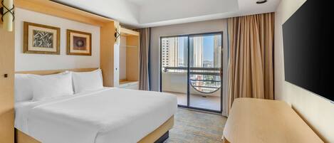 A hotel room with a large bed, a flat-screen TV, a balcony with a view of high-rise buildings, and a wooden headboard.