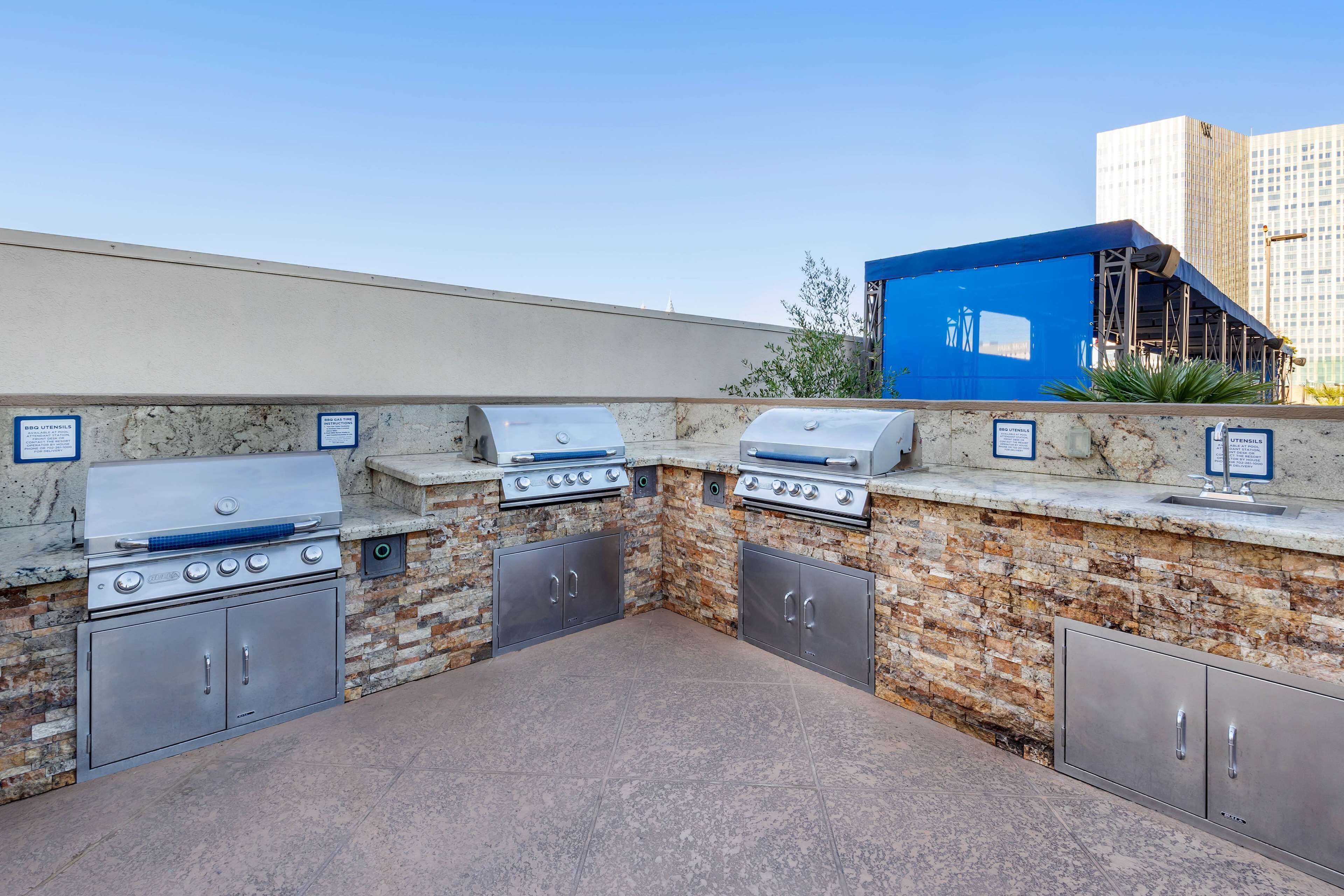 Una cocina exterior con dos parrillas, un lavamanos y armarios de almacenamiento, frente a una pared de piedra con vistas despejadas de un paisaje urbano.