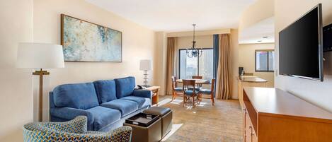 Quarto de hotel com sofá azul, poltrona estampada, mesa de jantar, lustre, televisão de tela plana e uma janela grande com cortinas.