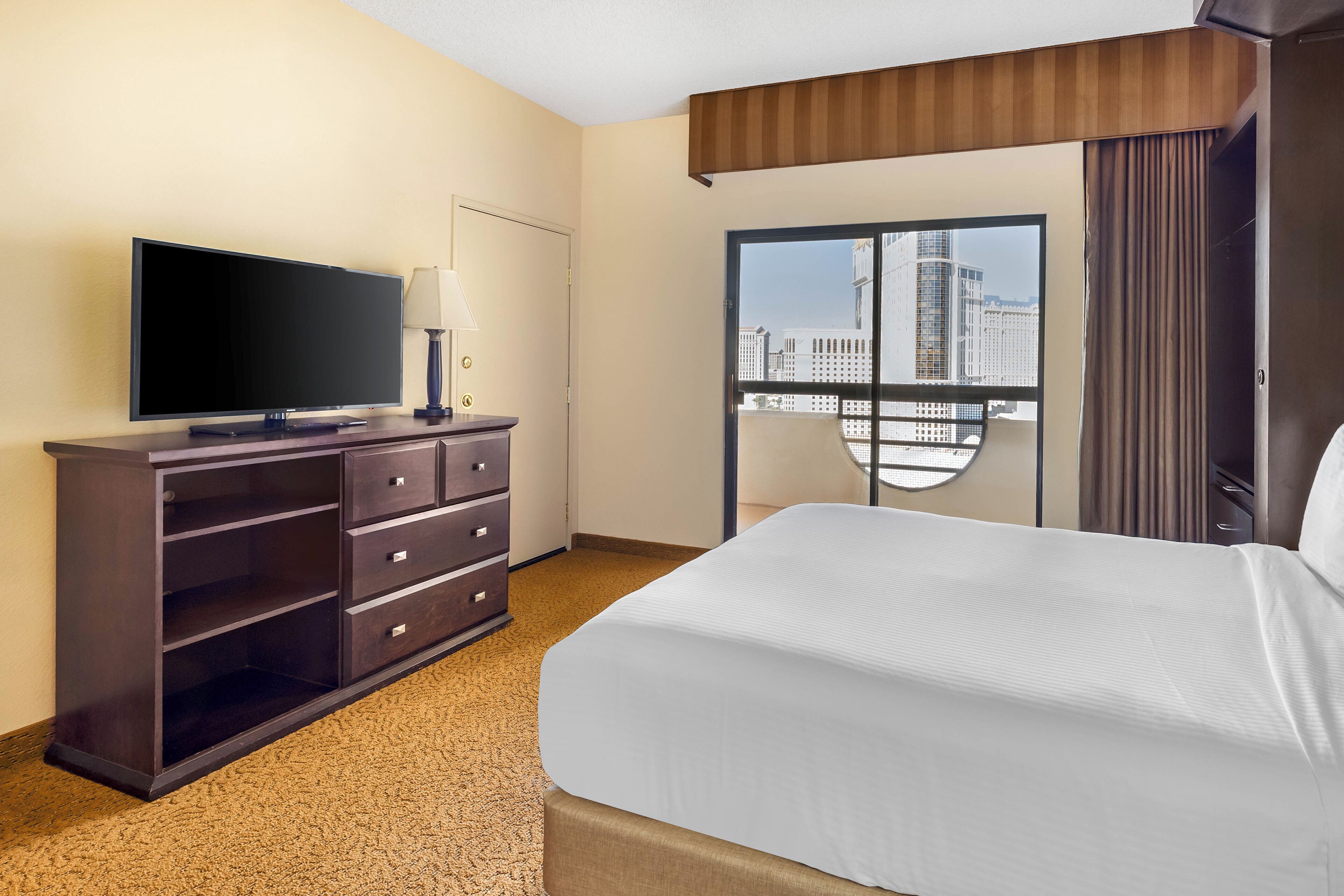 Habitación de hotel con una cama grande, un televisor de pantalla plana sobre un aparador de madera oscura, una lámpara y una ventana con vista a la ciudad.