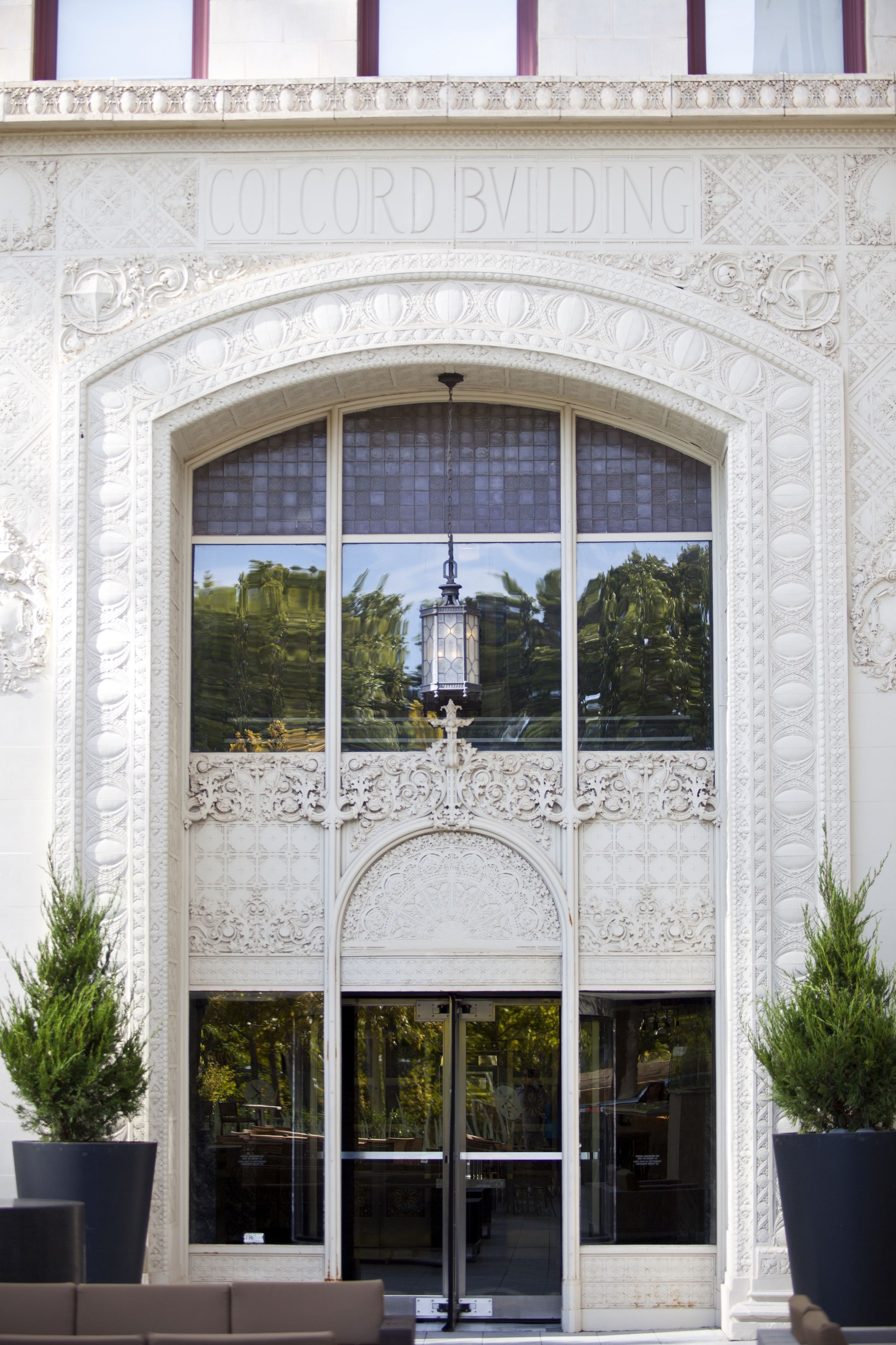 Exterior del edificio Colcord con detalles arquitectónicos ornamentados, amplios ventanales de vidrio y dos plantas en macetas en la entrada.