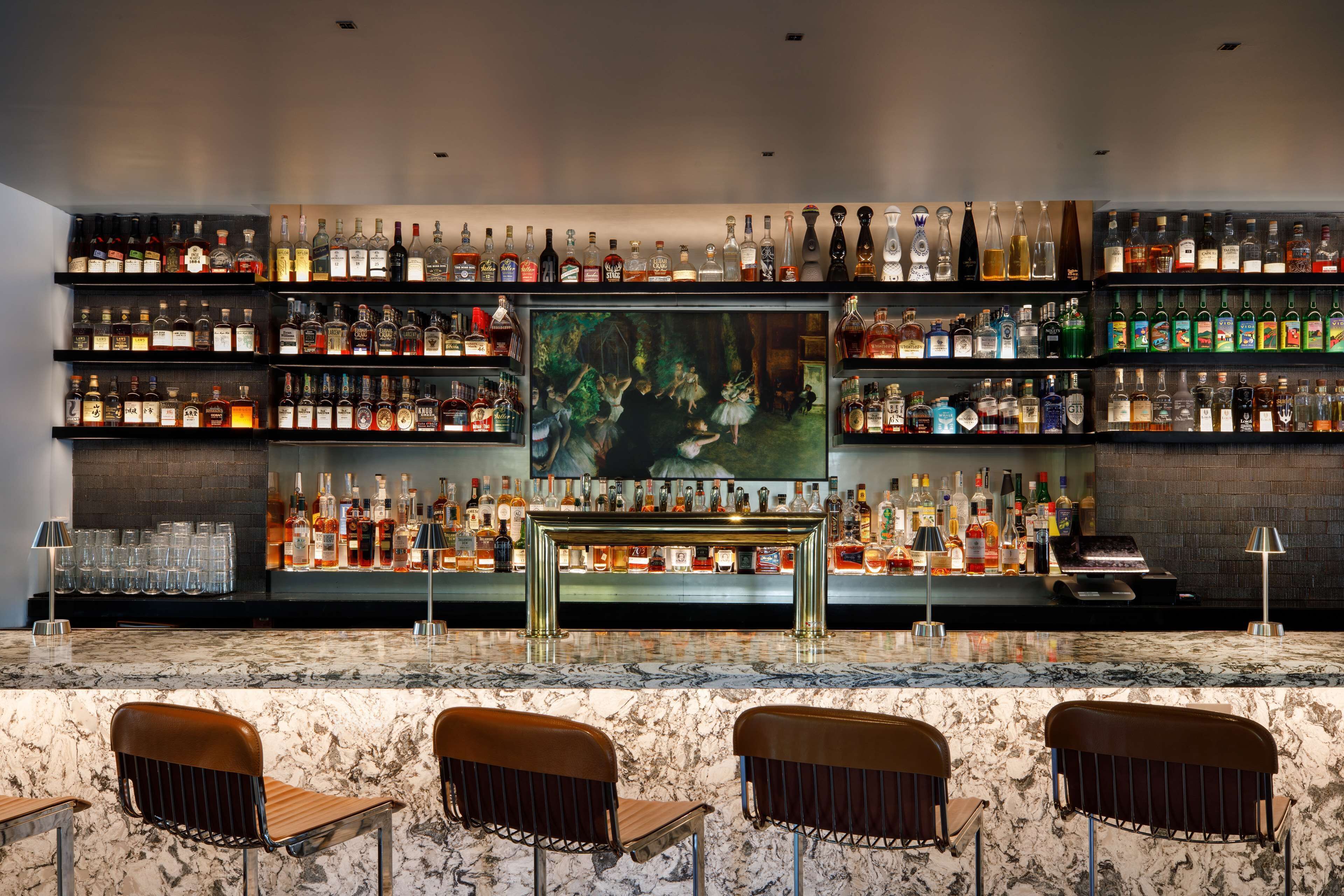 Un espace bar doté de comptoirs en marbre, de plusieurs étagères remplies de bouteilles d’alcool et de trois tabourets de bar.