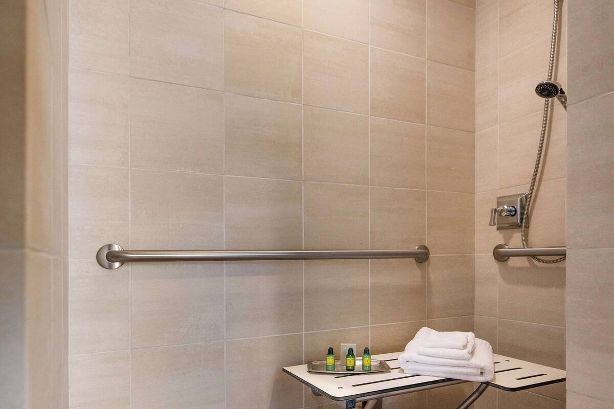 Un baño pequeño con azulejos color beige, una ducha, una barra de apoyo y un estante con artículos de aseo.