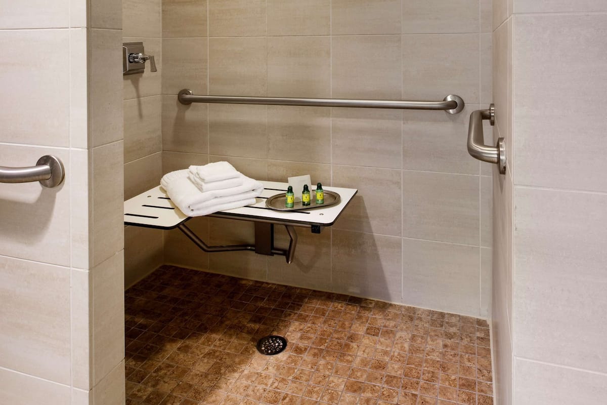 Un vestuario de ducha con azulejos color crema, una barra de apoyo, un perchero y un cabezal de ducha de mano.
