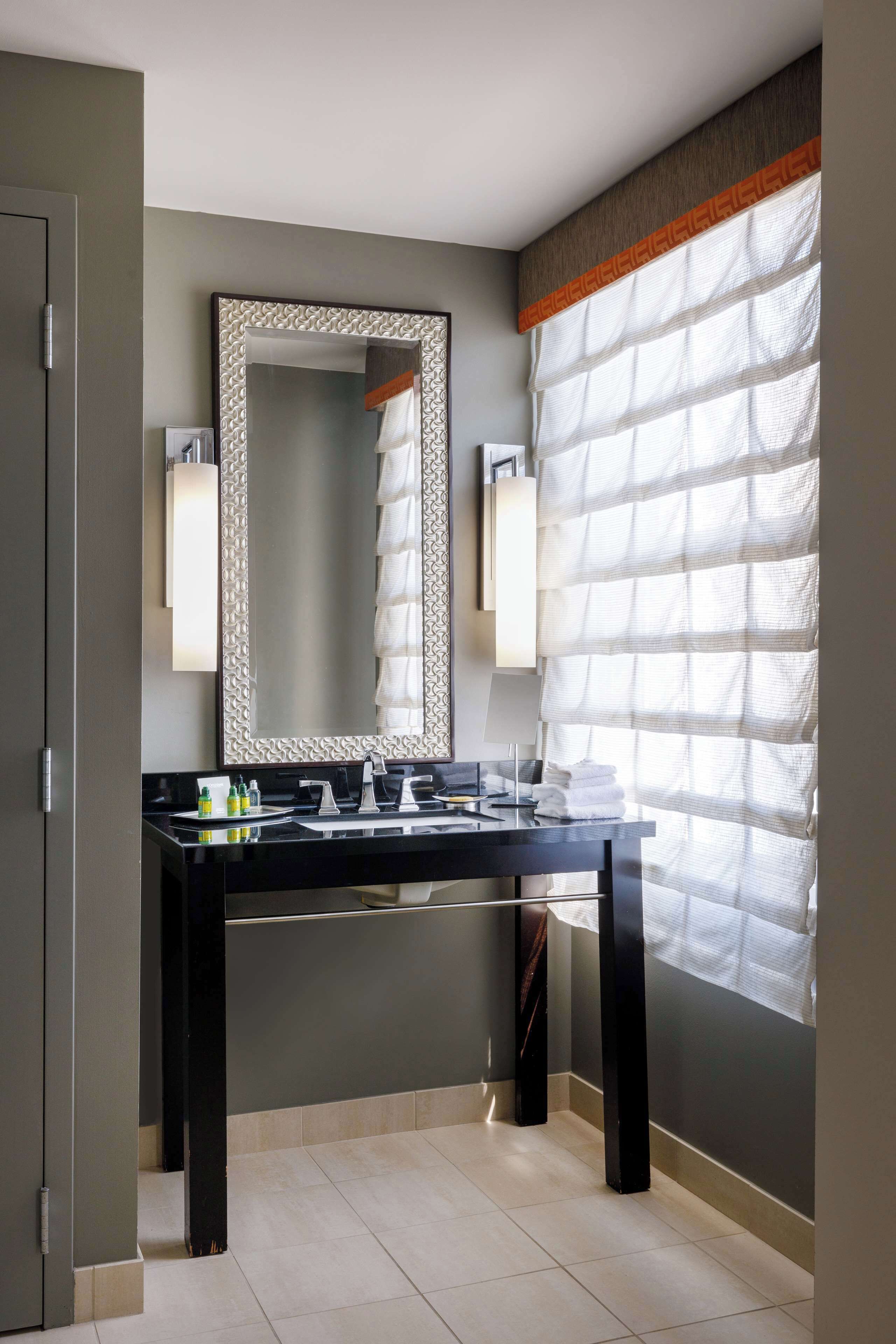 Une salle de bain moderne avec un grand miroir, un plan de toilette noir et une fenêtre ornée de rideaux légers.