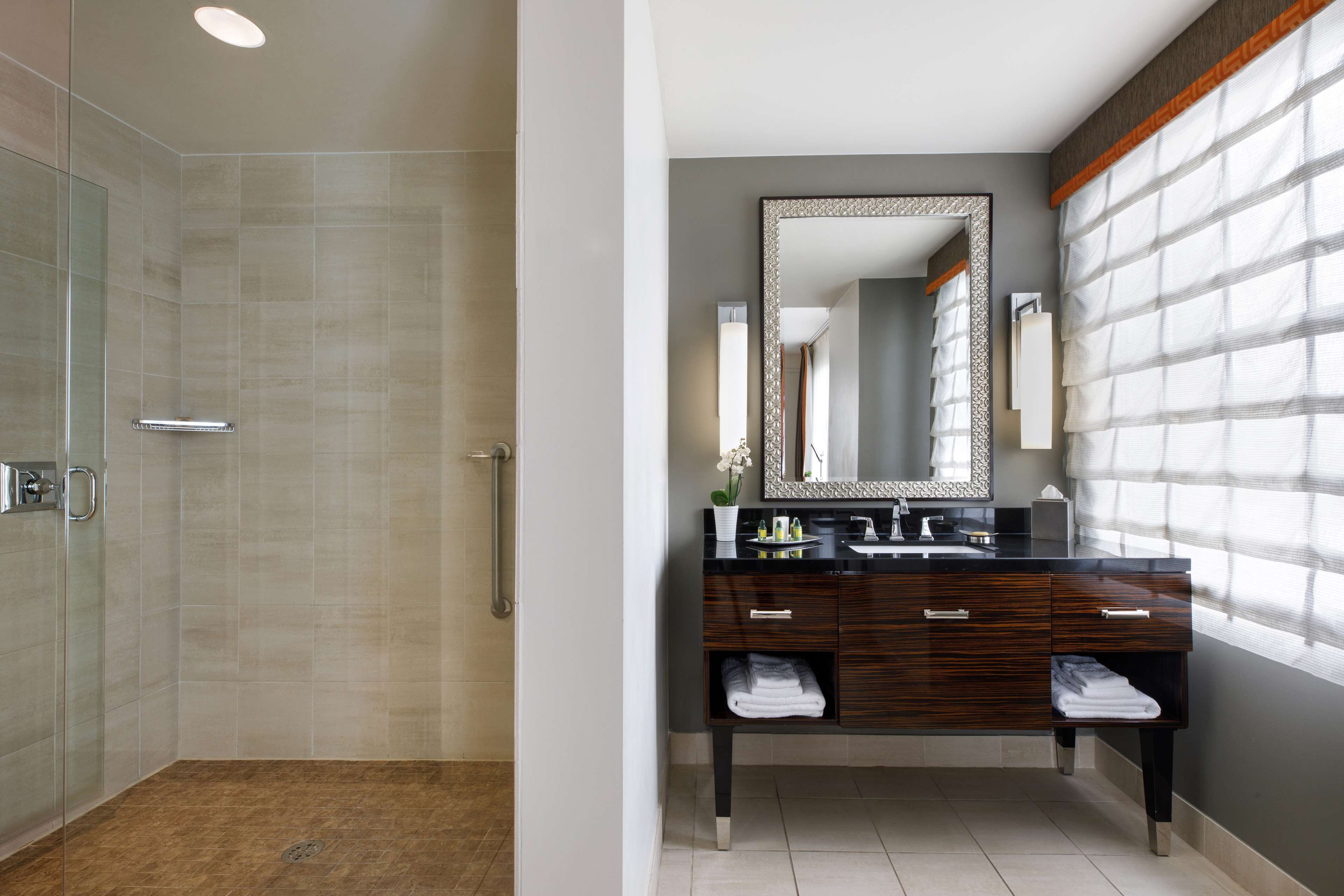 A modern bathroom with a glass-enclosed shower, a vanity with a large mirror, and a window with blinds.
