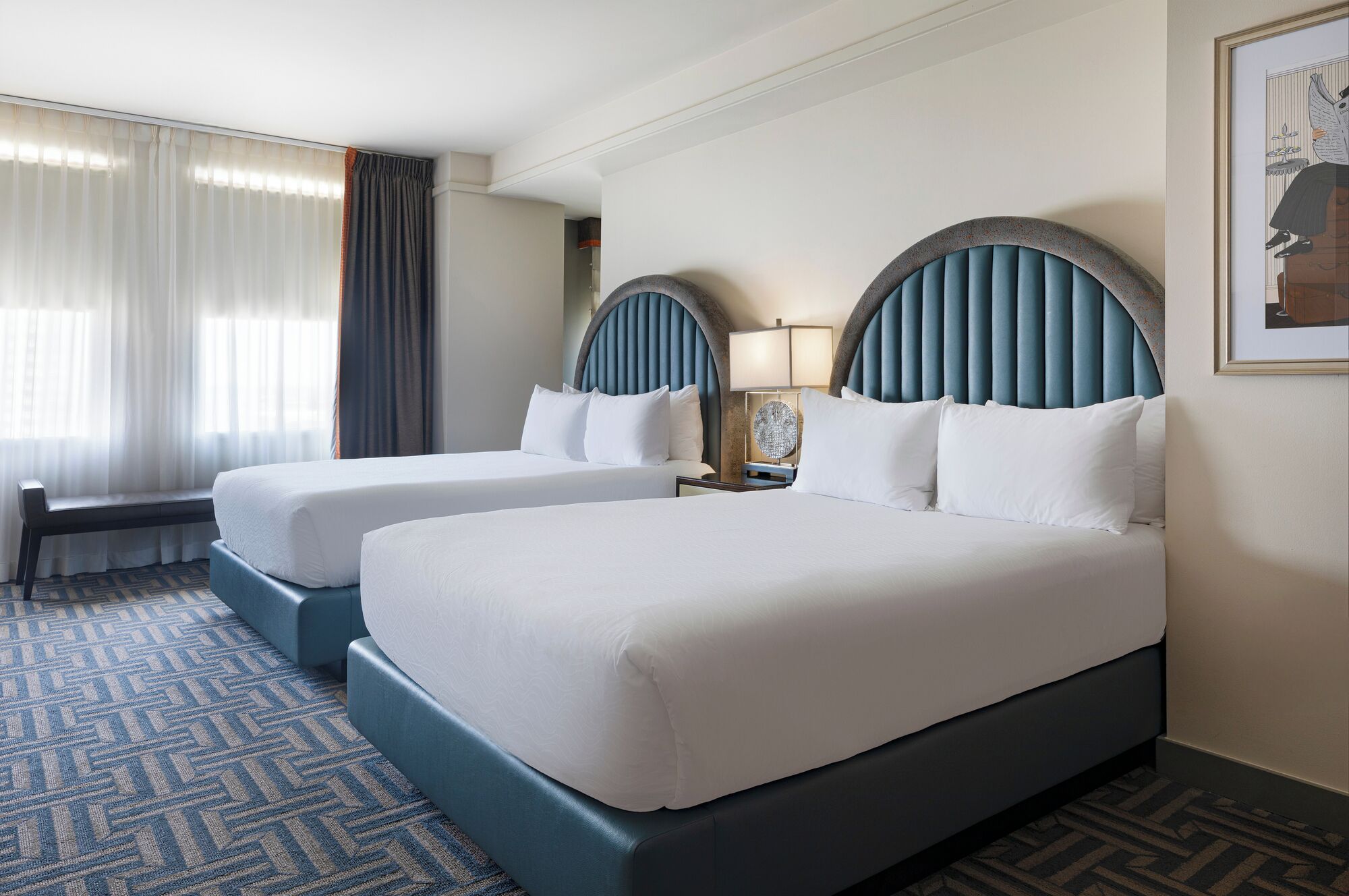 A hotel room with two beds, a bench, a nightstand with a lamp, and a framed picture on the wall.