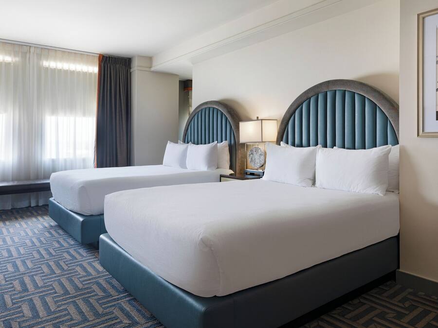 A hotel room with two beds, a bench, a nightstand with a lamp, and a framed picture on the wall.