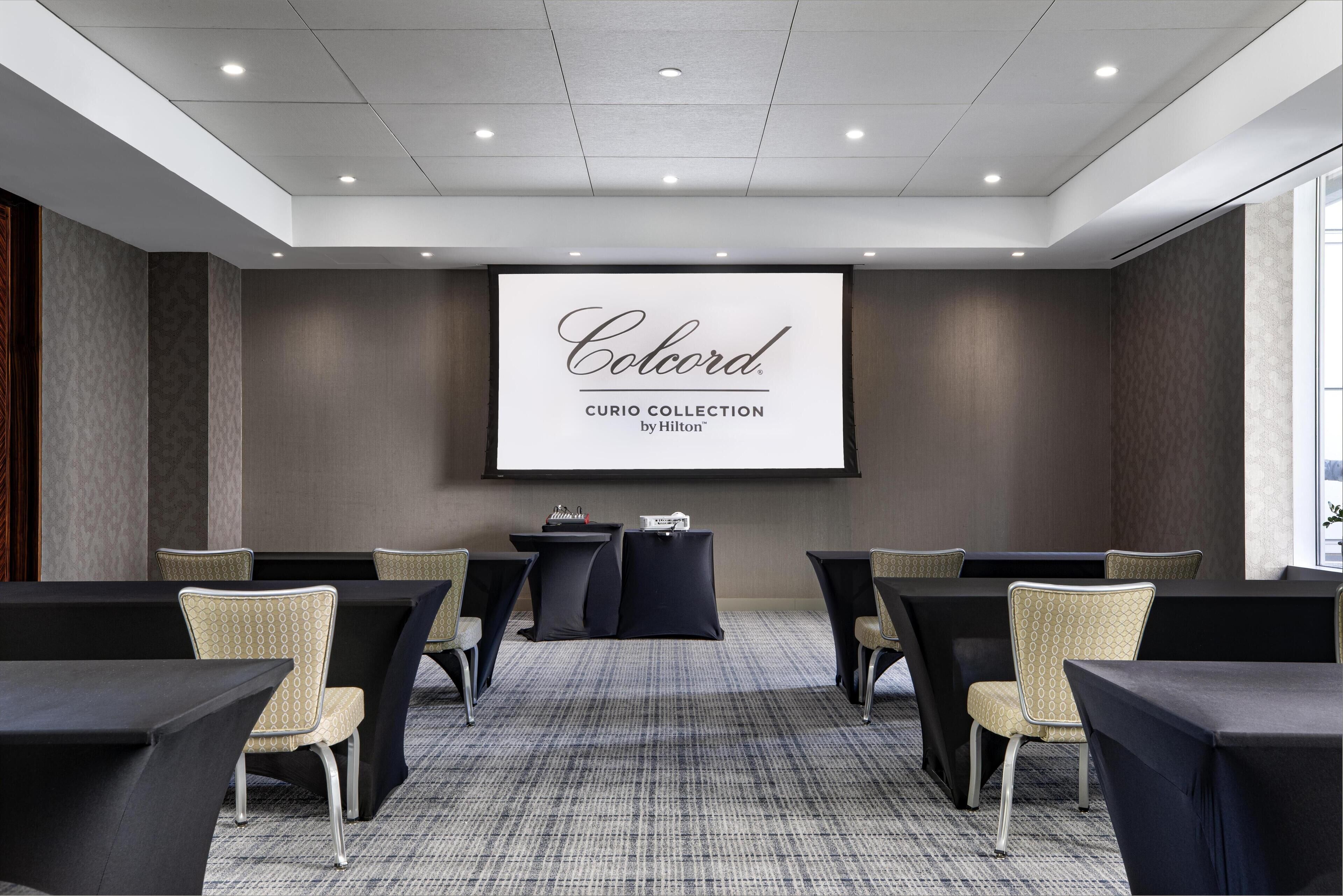 A conference room with a large screen displaying 'Colcord CURIO COLLECTION by Hilton', black tables, and wicker chairs.