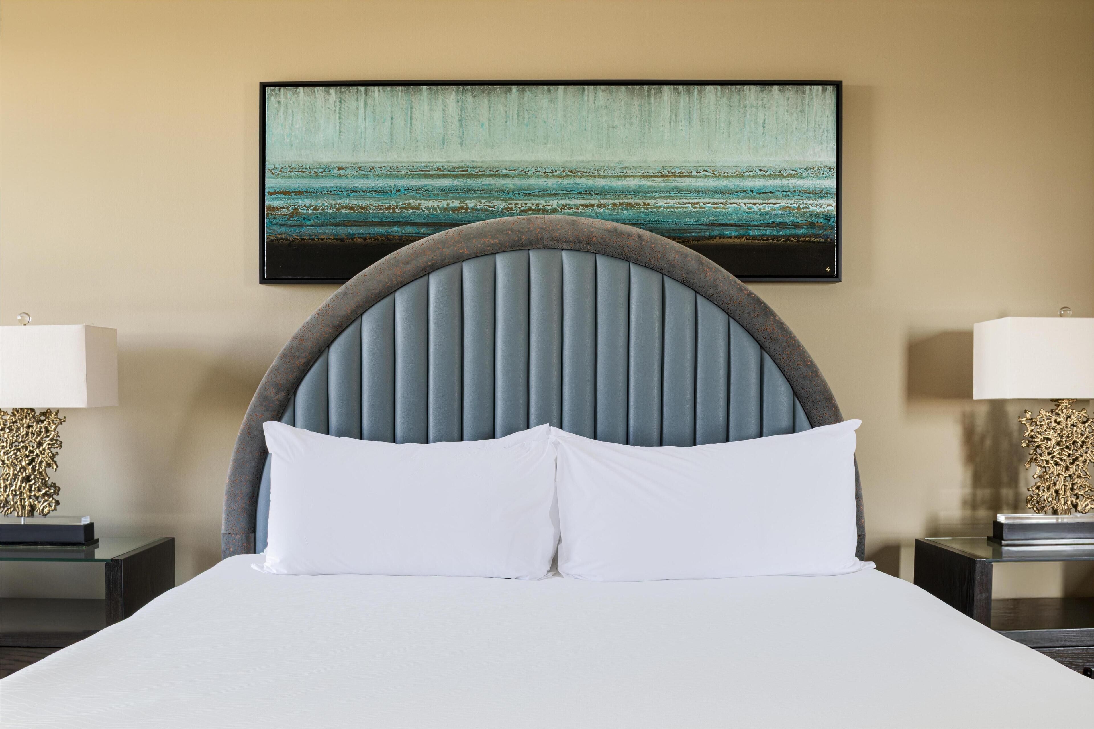 A hotel room with a large bed, a headboard with vertical slats, two bedside tables with lamps, and a painting of a coastal scene on the wall.
