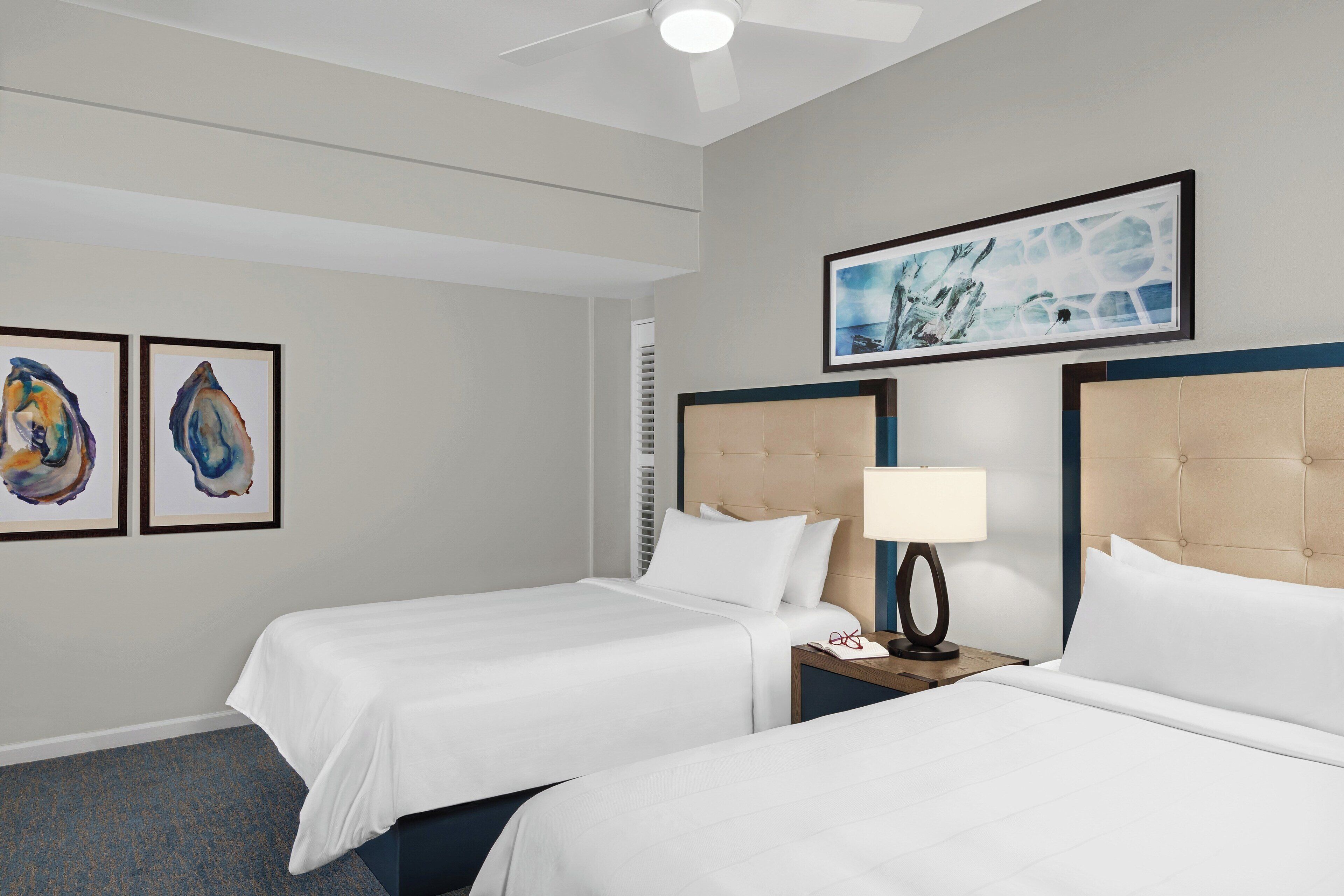 A hotel room with two beds, a ceiling fan, artwork on the walls, and a nightstand with a lamp.