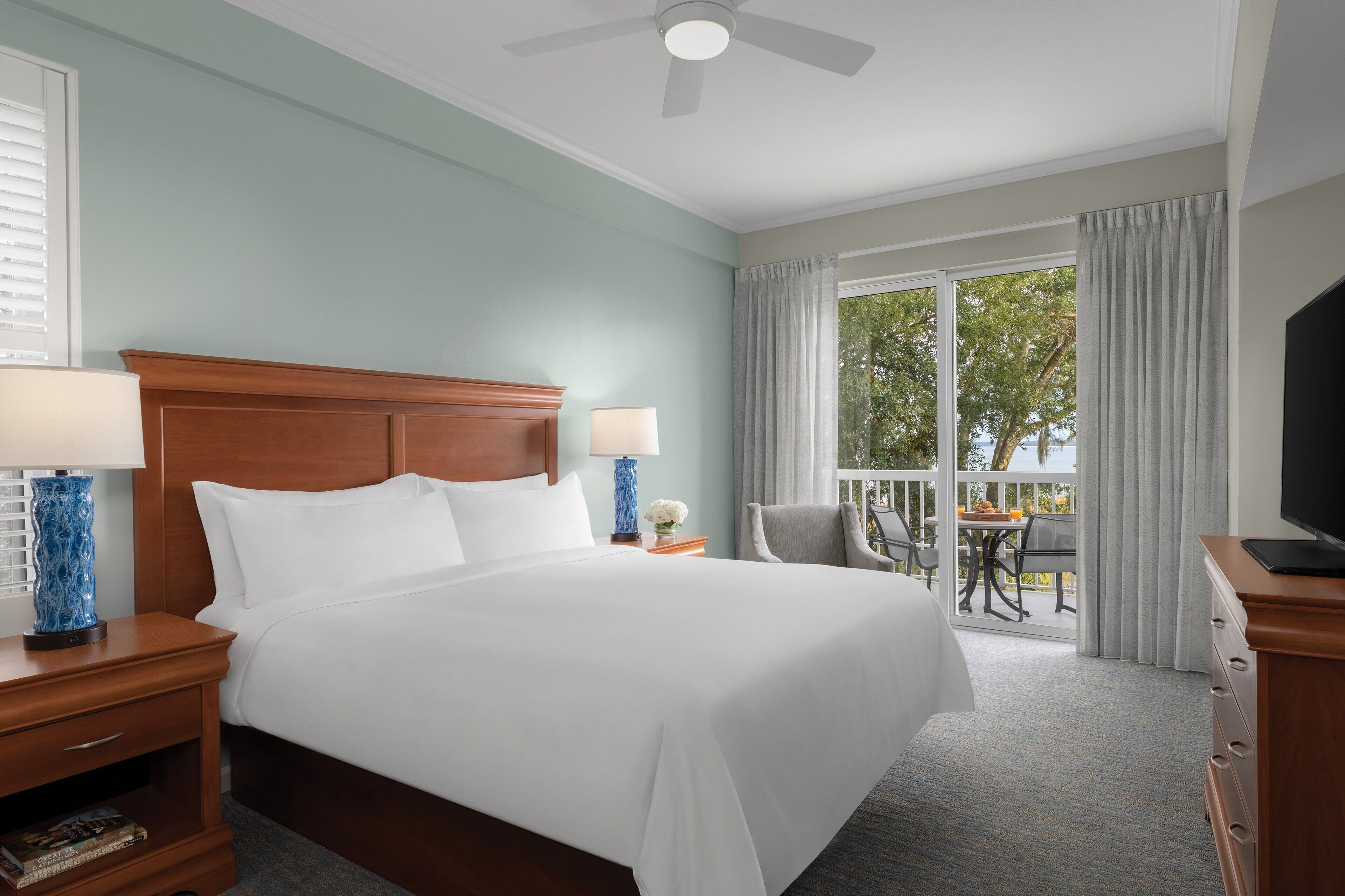 A hotel room with a large bed, two bedside tables with lamps, a TV, and a balcony with a table and chairs.