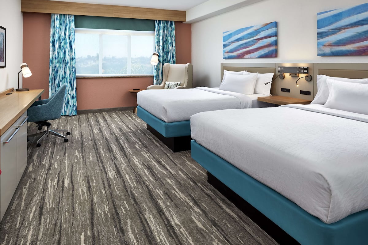 A hotel room with two beds, a desk, a chair, a window with curtains, and artwork on the walls.