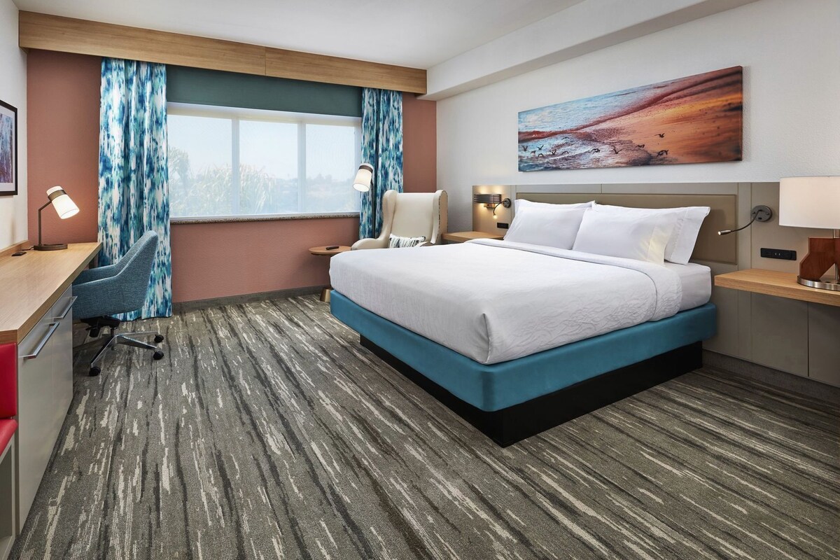 A hotel room with a large bed, a desk with a chair, a nightstand, a lamp, and a painting on the wall.