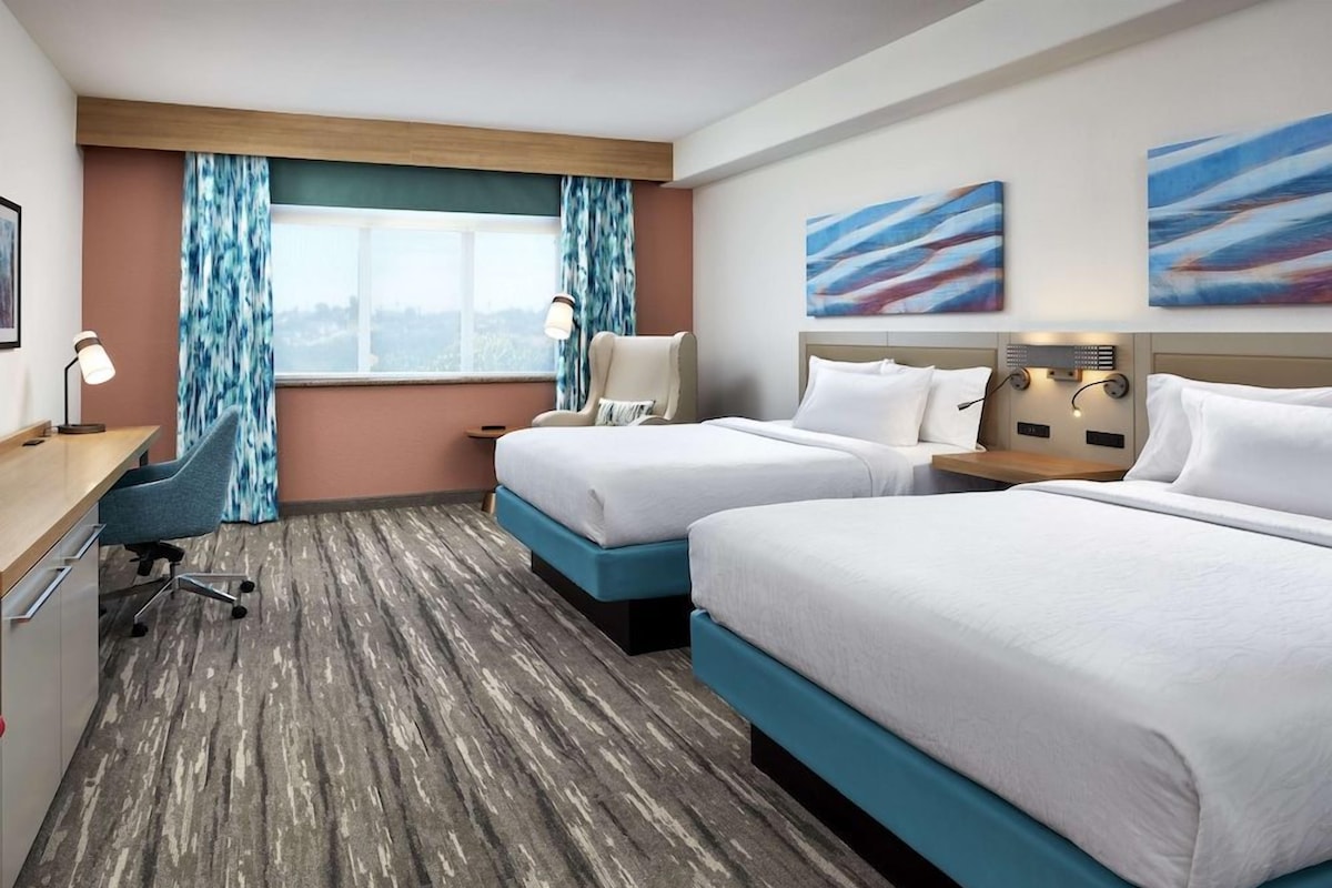 A hotel room with two beds, a desk, a chair, a window with curtains, and abstract wall art.
