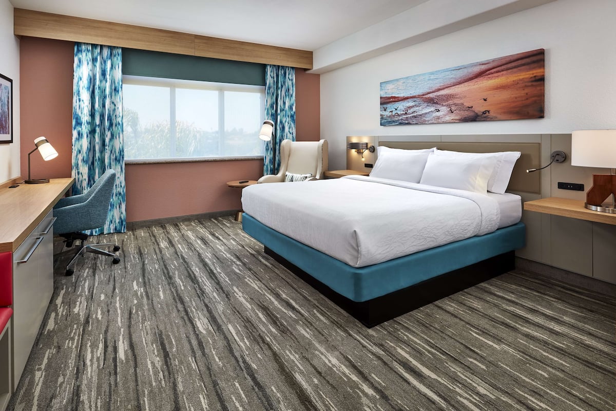 A hotel room with a large bed, a desk with a chair, a desk lamp, a nightstand with a lamp, and a painting on the wall.
