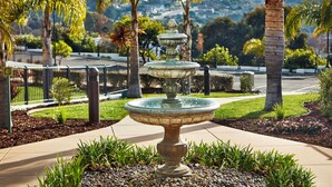 A multi-tiered fountain in a landscaped area with palm trees and a view of a hilly landscape in the background.