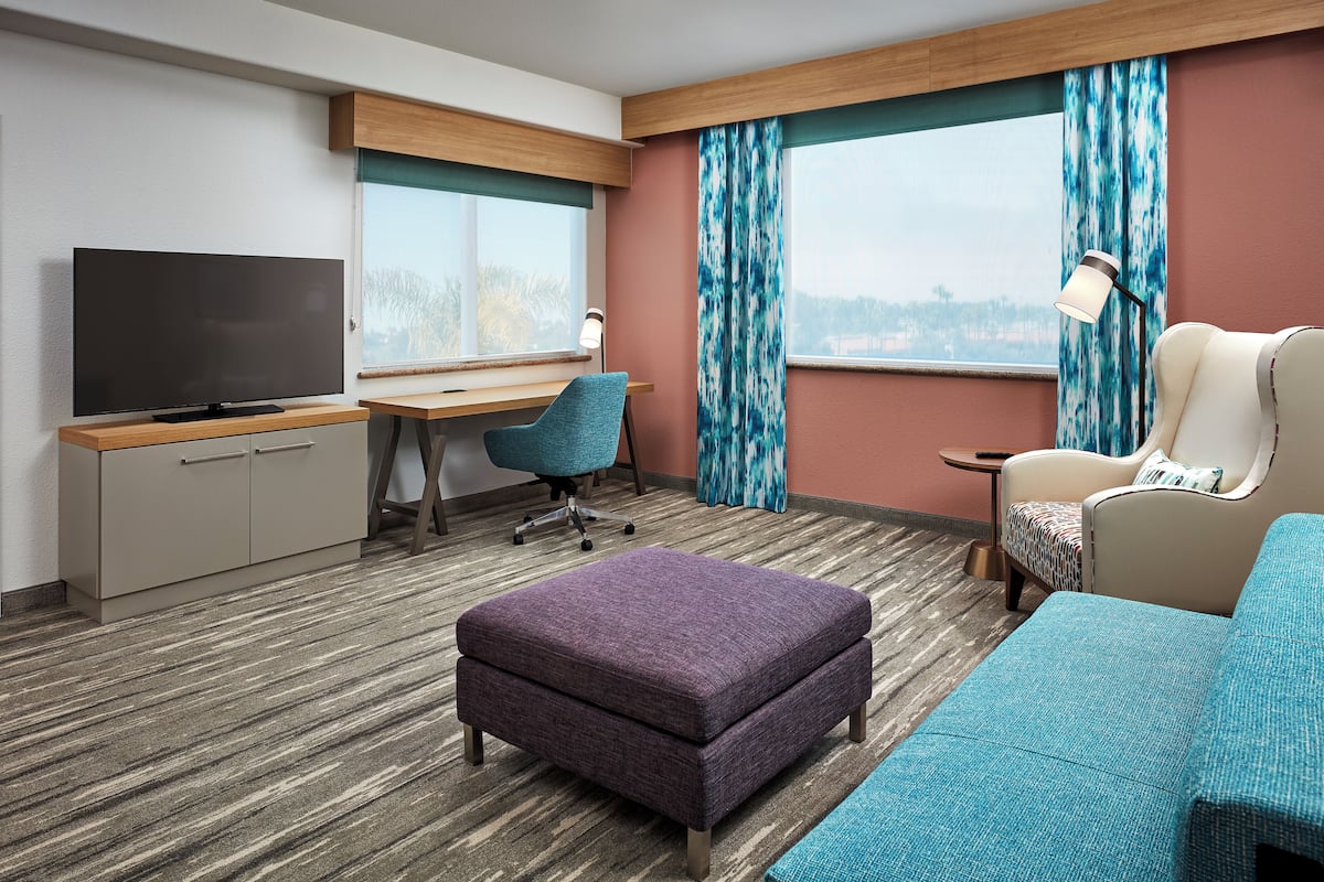 A hotel room with a flat-screen TV, a desk, a blue office chair, a purple ottoman, a teal sofa, and a window with curtains.
