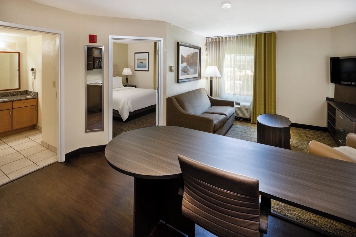 Habitación de hotel con zona de comedor, sofá, televisión y baño visible a través de una puerta abierta.