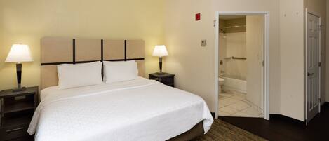 A hotel room with a large bed, two bedside tables with lamps, a bathroom with a bathtub, and a door leading to another room.
