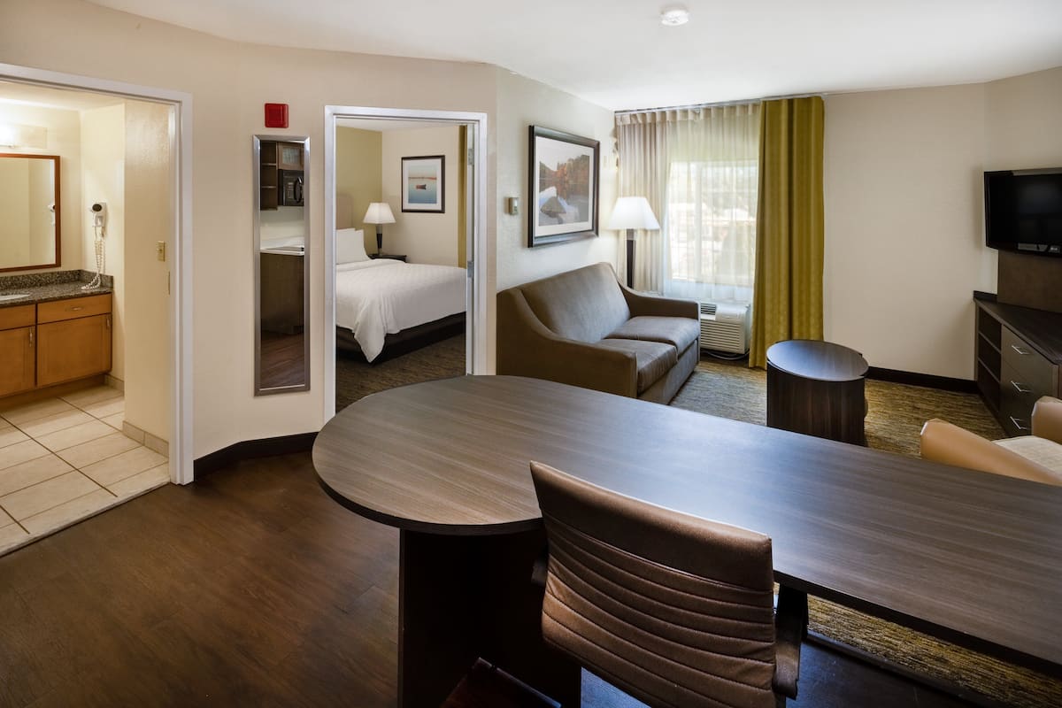 Habitación de hotel con zona de comedor, sofá, televisión y baño visible a través de una puerta abierta.