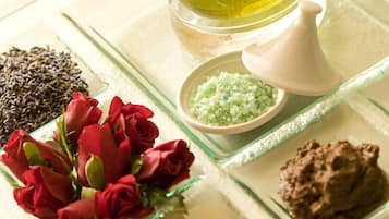 A glass tray with a bottle of oil, a bowl of green bath salts, a pile of black seeds, and a bouquet of red roses.