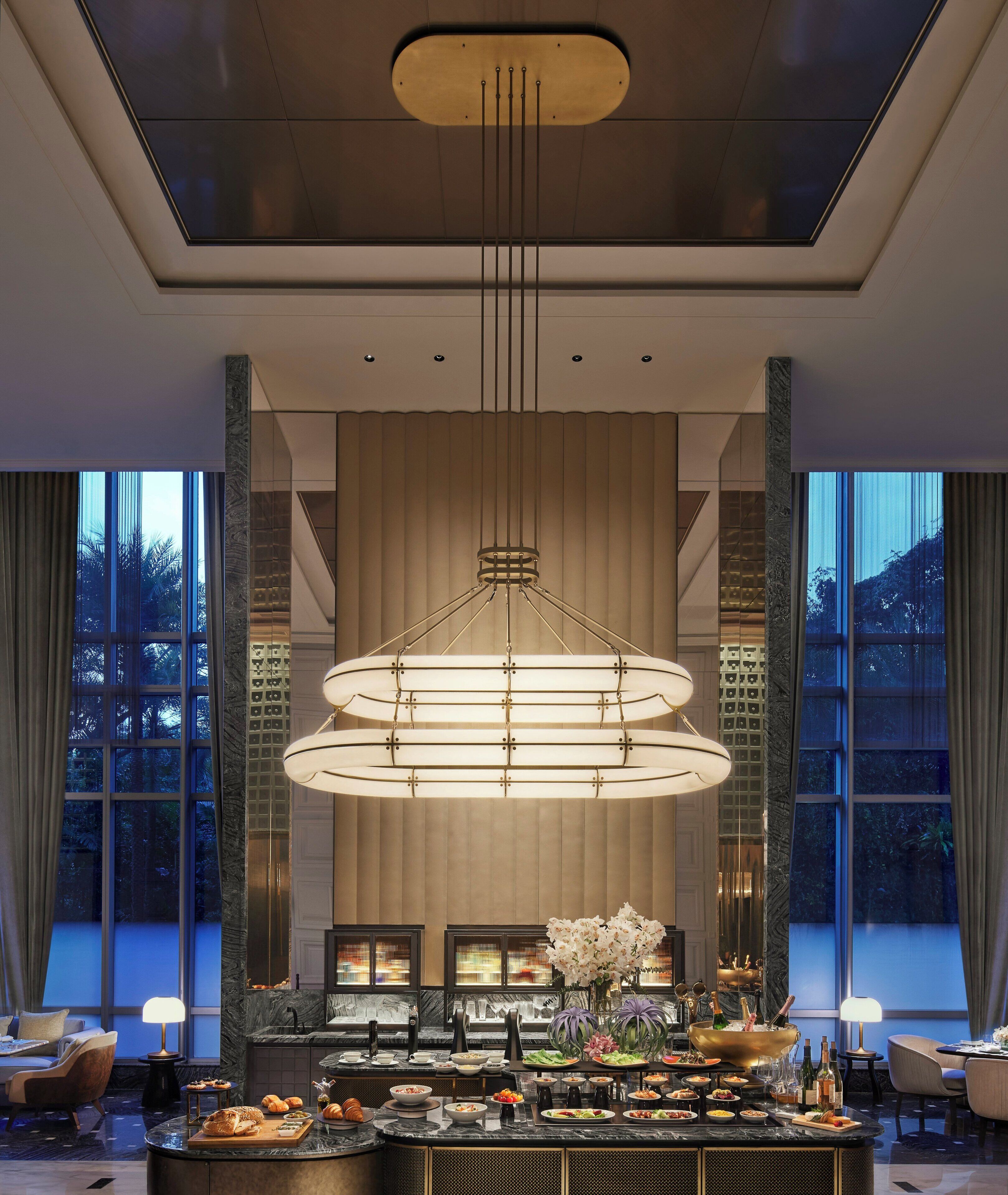 A modern hotel lobby with a central chandelier, a buffet area, and large windows offering a view of the outdoors.