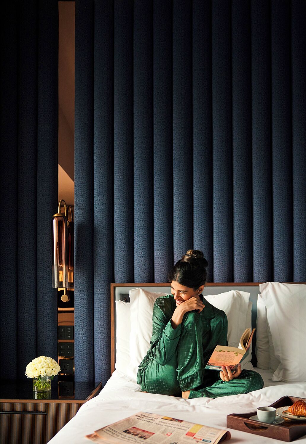 A person sitting on a bed in a room with a patterned blue curtain, a bedside table with a vase of flowers, and a tray with a croissant and a cup.