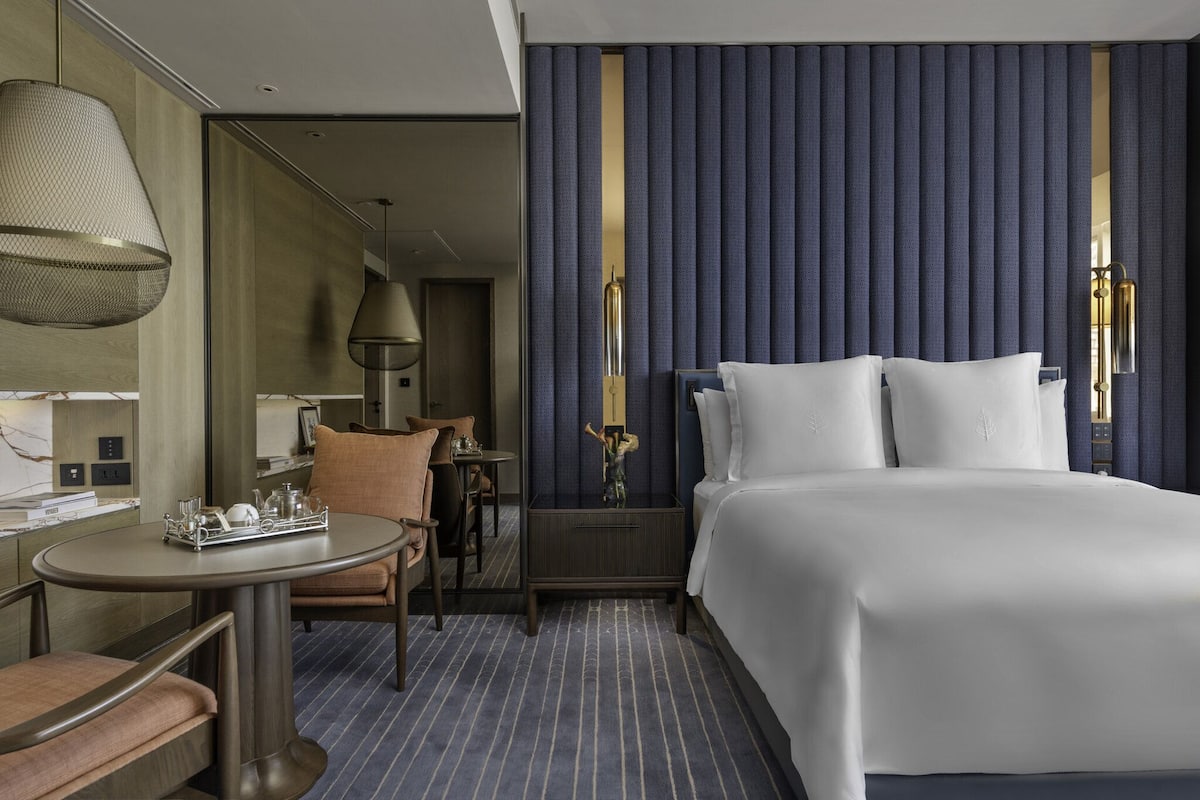 Ein Hotelzimmer mit einem großen Bett, einem runden Tisch mit Tablett und Gegenständen, einem Stuhl, einer Lampe und einer Wand mit vertikalen Jalousien.
