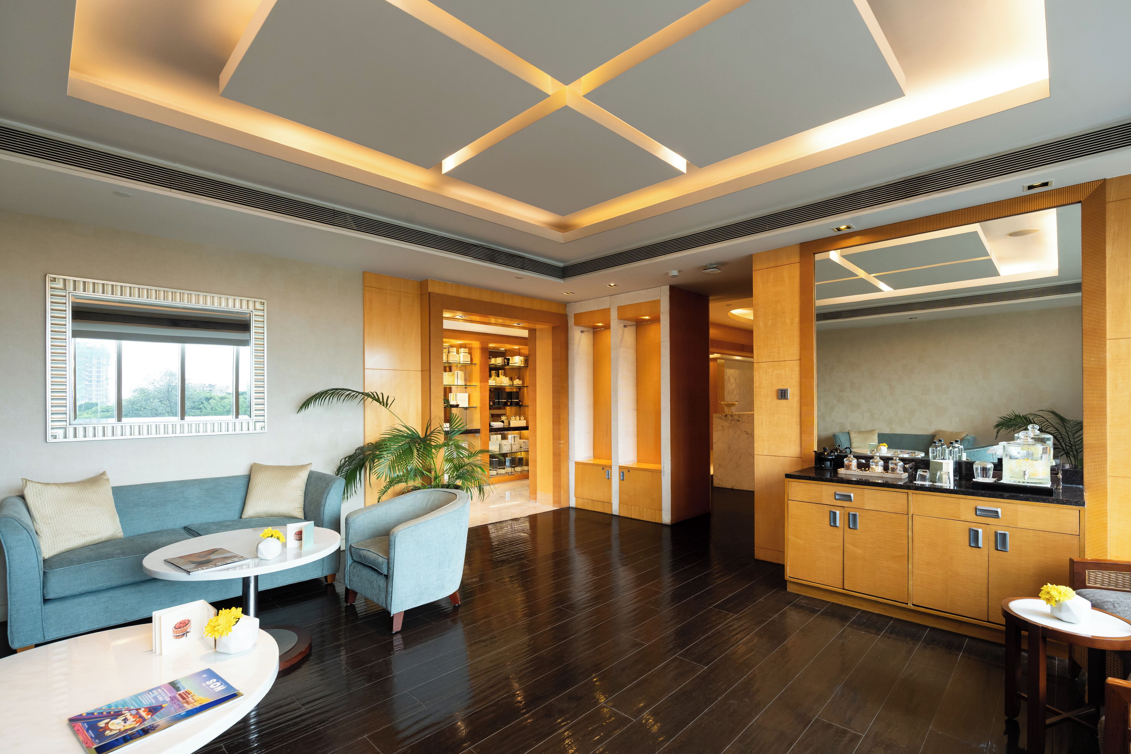 A modern hotel lobby with a blue sofa, a round coffee table, a seating area with a blue armchair, a kitchen area with wooden cabinets, and a large window with blinds.
