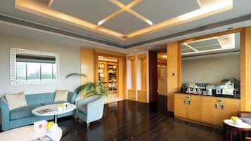 A modern hotel lobby with a blue sofa, a round coffee table, a seating area with a blue armchair, a kitchen area with wooden cabinets, and a large window with blinds.