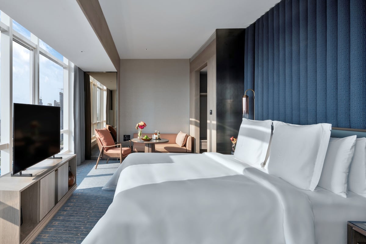 Ein modernes Hotelzimmer mit einem großen Bett, einem Fernseher, einer Sitzecke mit Tisch und großen Fenstern mit Blick auf die Stadt.