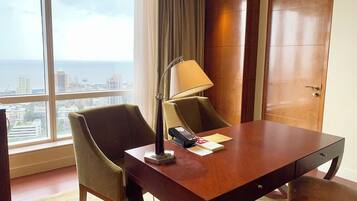 A hotel room with a large window, a desk with a lamp, a chair, and a wooden door.