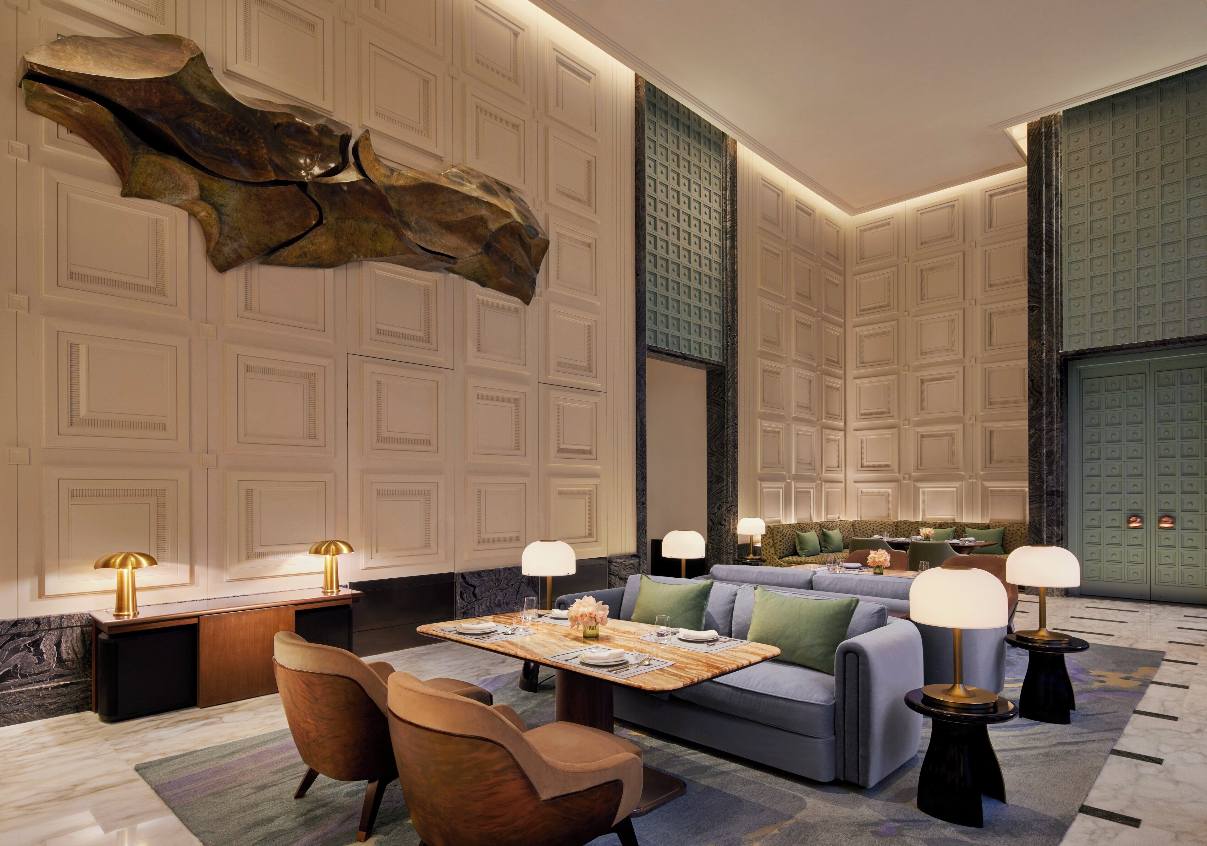 A modern hotel lobby with a marble floor, a large abstract sculpture, a seating area with sofas and armchairs, and a dining table set for two.