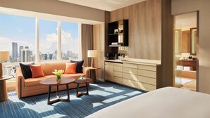Habitación de hotel moderna con una cama grande, un sofá, una mesa de centro y vistas de la silueta urbana a través de amplias ventanas.