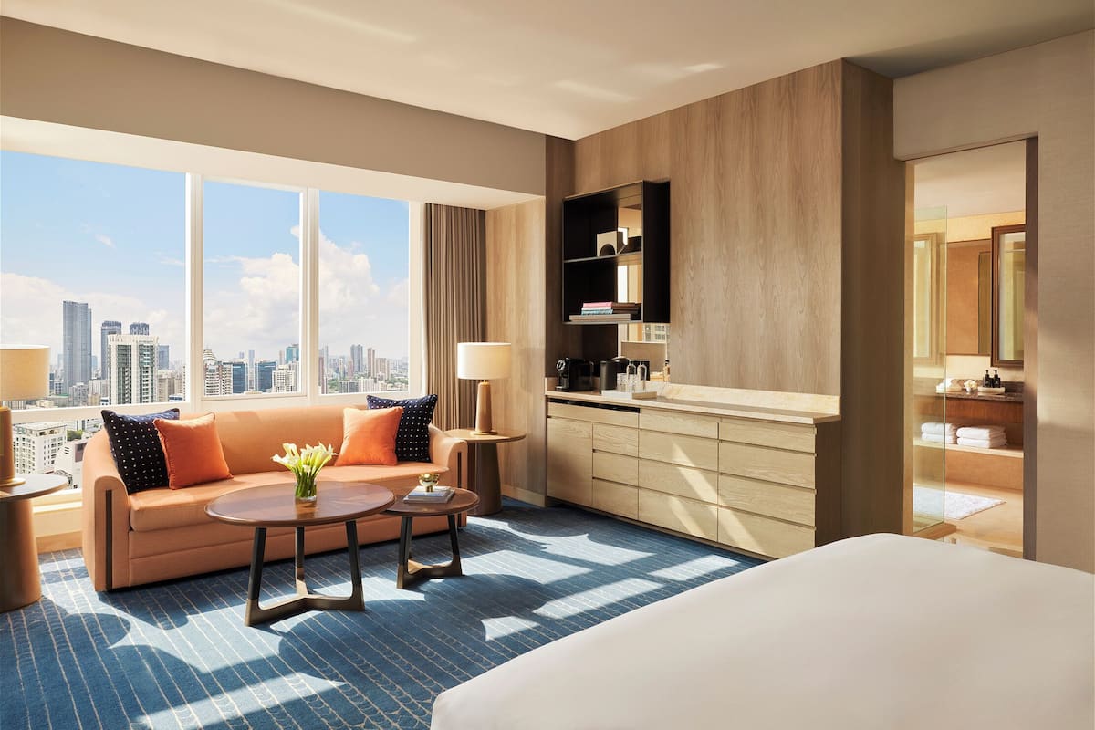 Ein modernes Hotelzimmer mit einem großen Bett, einer Couch, einem Couchtisch und Blick auf die Skyline der Stadt durch große Fenster.