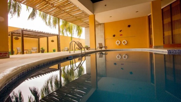 Piscina com borda revestida, área coberta com cadeiras e plantas em vasos, e parede amarela com detalhes decorativos.
