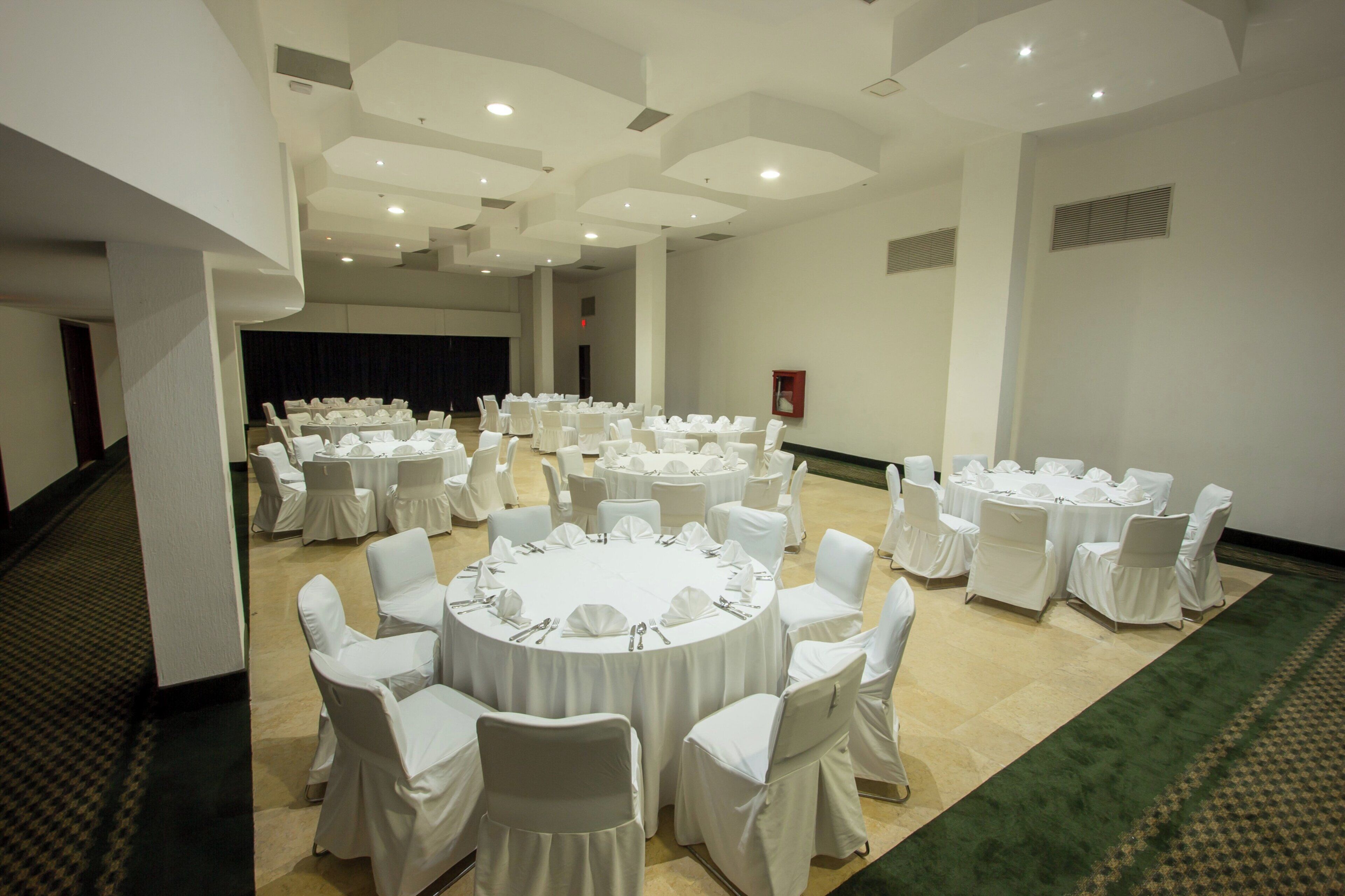 Un salón de conferencias amplio con varias mesas redondas, cada una rodeada de sillas cubiertas con manteles y fundas blancas.