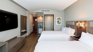 Quarto de hotel com televisão de tela plana, cómoda de madeira, cama com lençóis brancos, mesa de cabeceira e luminária fixada na parede.