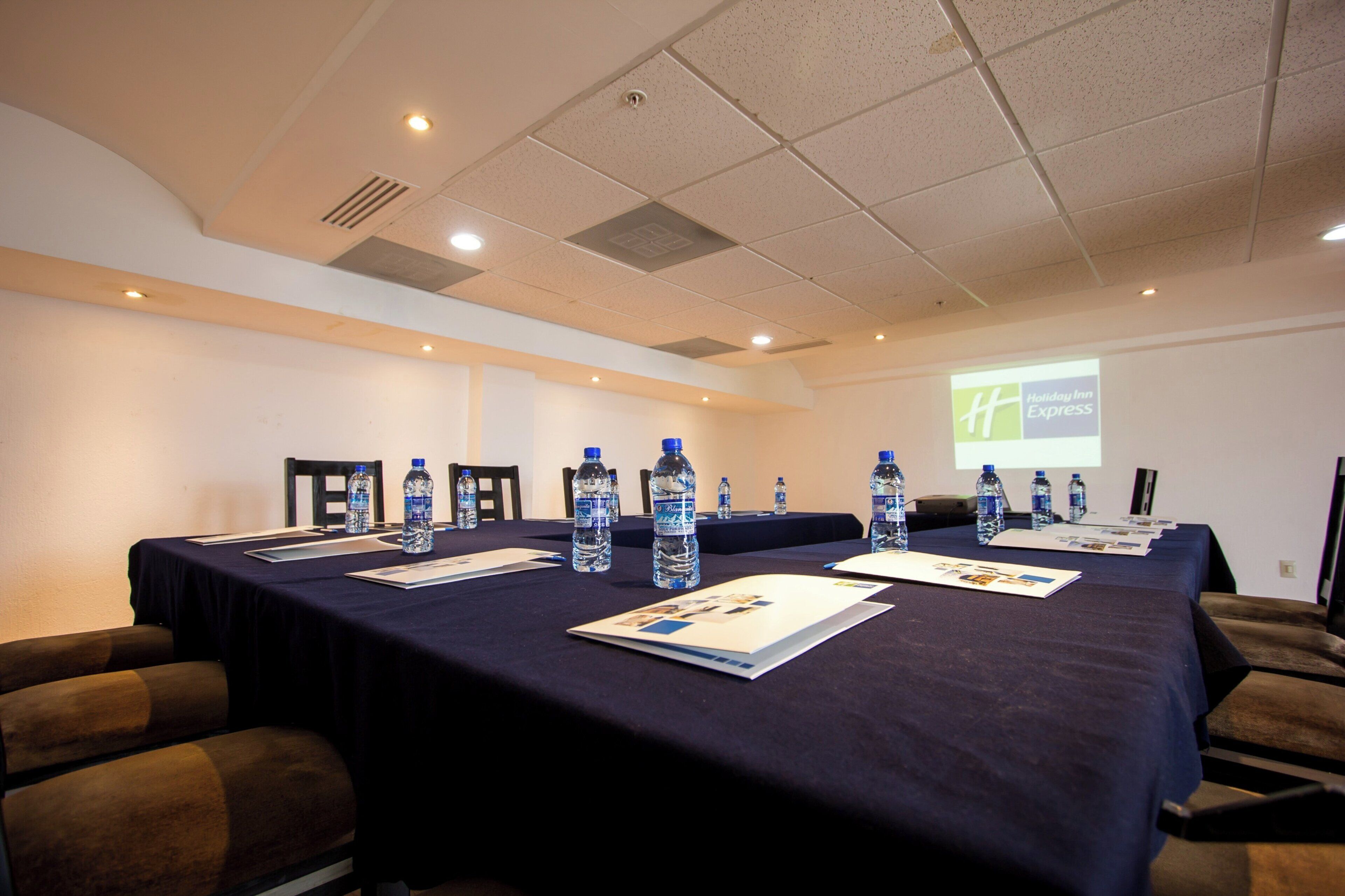 A conference room with a long table, chairs, water bottles, and a projector screen displaying the Holiday Inn Express logo.