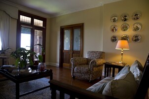 Una sala de estar con sofá, sillón, mesa de centro y una lámpara. Hay platos decorativos en la pared y una puerta con paneles de vidrio.