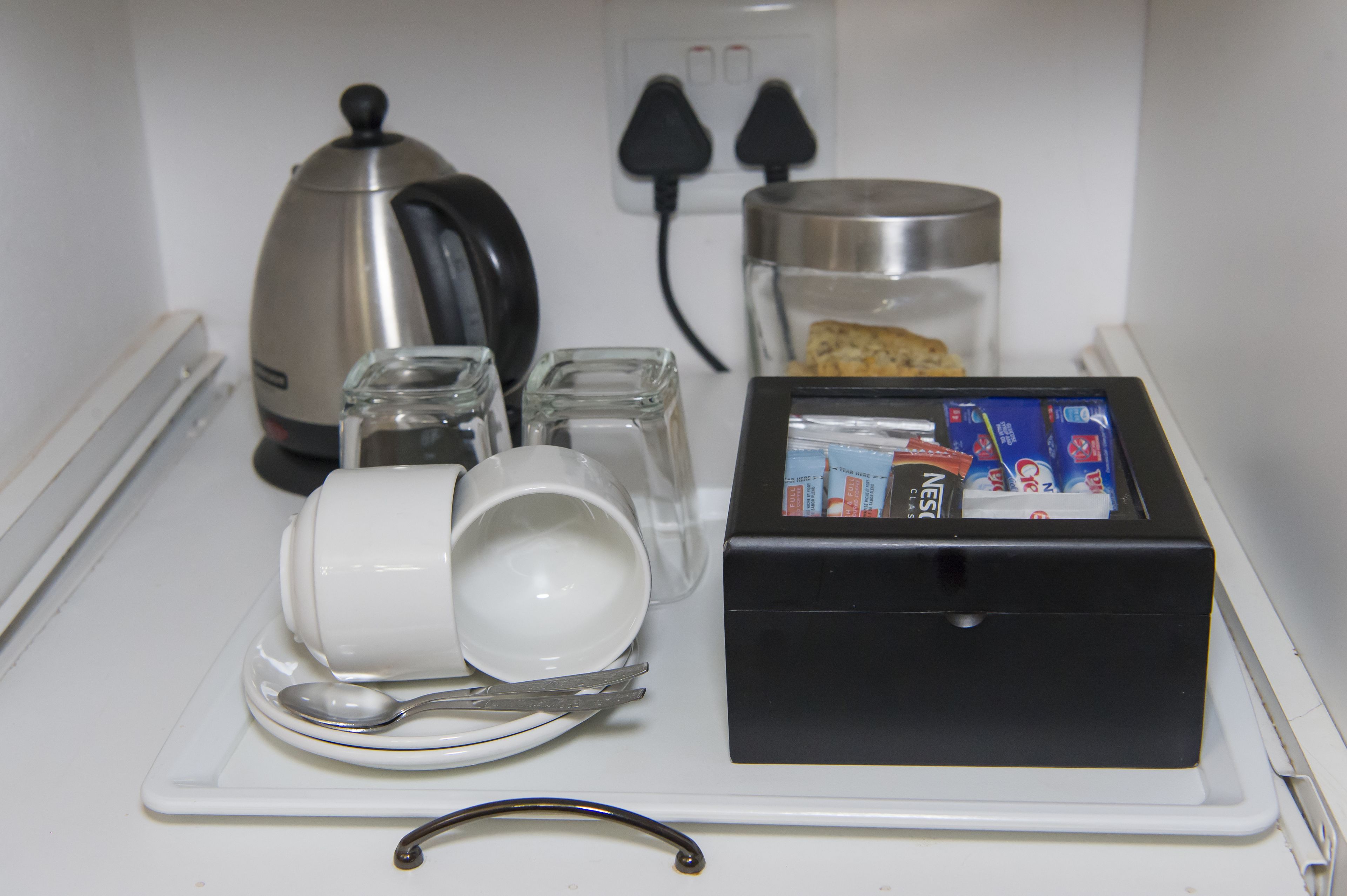 A kitchen counter with a coffee maker, a glass jar, a box of Nespresso coffee capsules, a white mug, a spoon, and a glass.