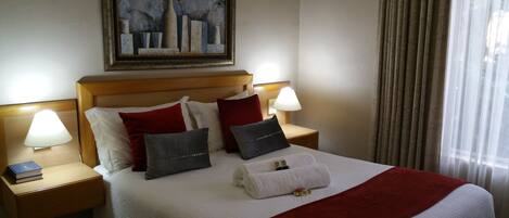 A neatly made bed with a red blanket, white pillows, and a painting above it. There are two bedside tables with lamps and a book on one.